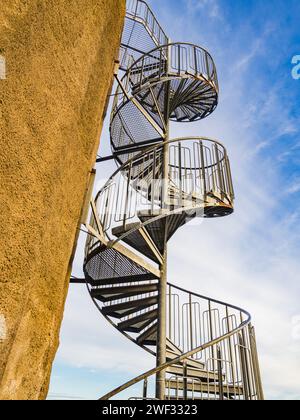 Questa immagine cattura l'intricato design di una scala a chiocciola in metallo che sale lungo l'esterno di un edificio beige sotto un cielo azzurro. Foto Stock