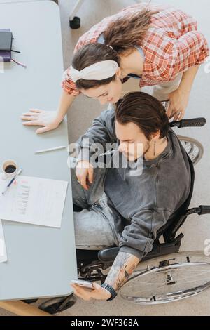 Una giovane donna premurosa si appoggia per assistere un uomo concentrato su una sedia a rotelle, che sta esaminando i grafici aziendali. La scena mette in evidenza l'inclusività e il lavoro di squadra Foto Stock