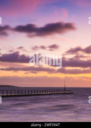 L'immagine cattura una serena scena del tramonto con un molo di legno che si estende nell'oceano sotto un cielo colorato. Foto Stock