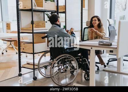 Donna di successo che discute di lavoro con il suo collega uomo handicappato in sedia a rotelle presso un moderno ufficio creativo. Foto Stock