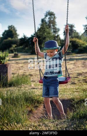 Un bambino con un cappello blu sta cercando di raggiungere e scalare l'altalena all'aperto da solo. Foto Stock