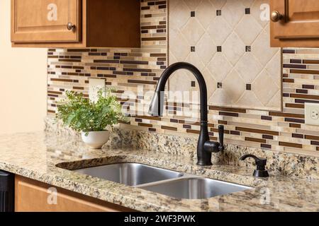 Un rubinetto da cucina con armadietti in legno, un rubinetto in bronzo strofinato a olio, un ripiano in quarzo e un backsplash in vetro e pietra. Foto Stock