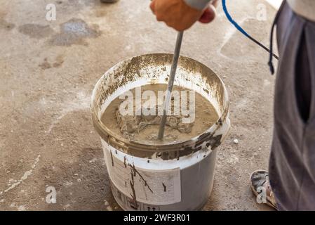 Il lavoratore mescola la soluzione in un secchio utilizzando un miscelatore. Miscelare la soluzione di intonaco in una benna utilizzando un trapano elettrico. Foto Stock
