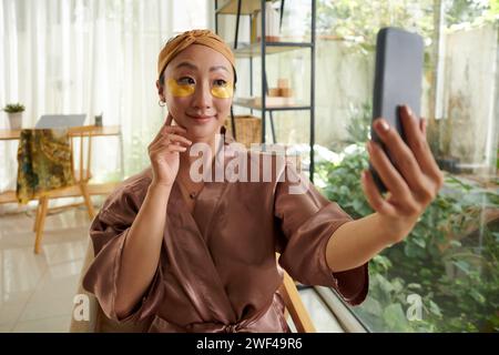 Ritratto di una giovane donna sorridente con cerotti sotto gli occhi che scattano selfie per il blog di bellezza Foto Stock