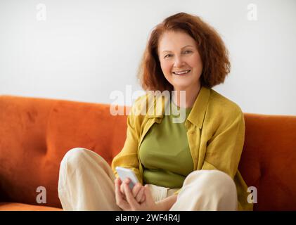 Donna anziana allegra con lo smartphone in mani sedute a gambe incrociate su un divano arancione Foto Stock