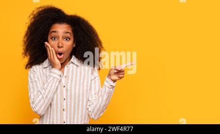 Donna nera scioccata che punta da un lato su sfondo giallo Foto Stock