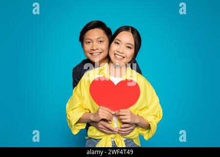 Una coppia radiosa si abbraccia con gioia, tenendo insieme un grande cuore rosso, con sorrisi luminosi e amore Foto Stock