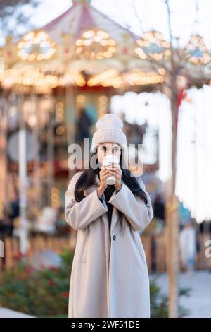 Giovane donna che si gode un drink caldo davanti a una giostra adornata di luci in una fredda giornata invernale, evocando un'atmosfera festosa. Foto Stock