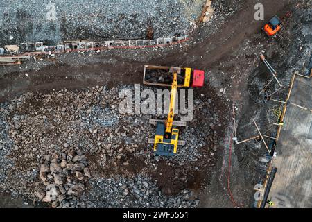 Vista aerea dall'alto verso il basso del cantiere con escavatore e dumper durante i lavori di terra. Scavatrice che carica ghiaia nella parte posteriore di un camion Foto Stock