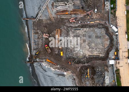 Vista aerea dall'alto verso il basso del cantiere con escavatori e dumper durante i lavori di terra e la costruzione di fondamenta di edifici sul lato del mare Foto Stock