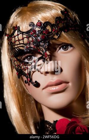 Manichino donna in plastica con capelli biondi luminosi che indossa una mezza maschera metallizzata nera con gemme rosse e abiti e accessori romantici che si posa su un bla Foto Stock