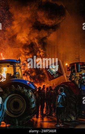 Turnhout, Anversa, Belgio, 28 gennaio 2024, questa potente immagine cattura un momento di protesta sotto il cielo notturno a Turnhout, in Belgio, dove gli agricoltori si sono riuniti per esprimere il loro dissenso contro l'accordo sull'azoto. Le braci brucianti e il fumo che si innalza nell'aria creano uno sfondo drammatico per i trattori illuminati di blu che fiancheggiano la scena. I manifestanti, visibili solo in sagoma, sono riuniti intorno al fuoco, con le fiamme che riflettono la loro determinazione e la gravità della loro causa. L'umore è intenso e carico, riflettendo le conversazioni cruciali intorno ad Agricult Foto Stock