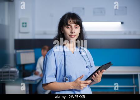 Potrait scatto di una donna operaia medica che detiene quello che sembra essere un tablet elettronico. Si trova in uno studio medico che guarda la macchina fotografica, mentre il medico maschile è sullo sfondo, Foto Stock