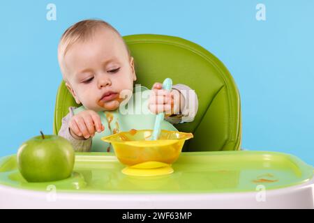 Piccolo bambino carino che mangia cibo sano in seggiolone su sfondo azzurro Foto Stock