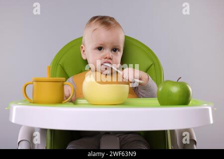 Piccolo bambino carino che mangia cibo sano in seggiolone su sfondo grigio Foto Stock