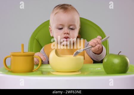 Piccolo bambino carino che mangia cibo sano in seggiolone su sfondo grigio Foto Stock