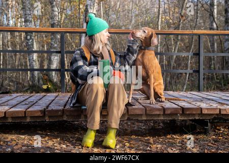 Escursionista stanco, amante degli animali domestici che riposa sul sentiero naturale dopo aver camminato nei boschi, tenendo in mano il thermos, accarezzando il cane Foto Stock