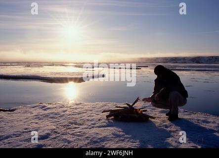 Uomo vicino a un incendio sul ghiaccio durante la rottura primaverile, sole di mezzanotte con nebbia che si snoda nella costa artica centrale dell'Alaska Foto Stock