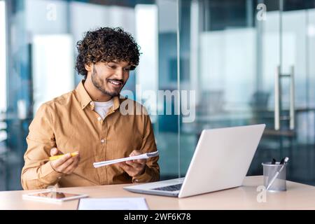 Uomo arabo attento che tiene un blocco note e una penna mentre si siede accanto al tavolo con un computer portatile in un luogo di lavoro moderno. Freelance positivi che scrivono i desideri e le idee dei clienti sulla realizzazione dei progetti tramite videochiamata. Foto Stock