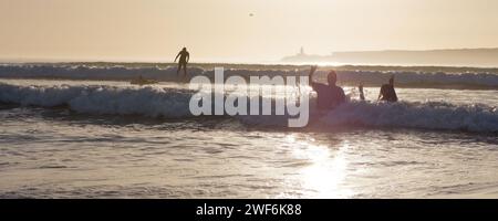 Surfista in silhouette cavalca un'onda al tramonto con persone in mare che ondeggiano in primo piano con un'isola alle spalle a Essaouira, Marocco, 28 gennaio 2024 Foto Stock