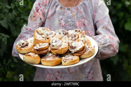 Piatto di panini nelle mani di una nonna dai capelli grigi felice, anziana nonna cuoce panini alla cannella e prepara biscotti. Verticale, focale selettiva Foto Stock