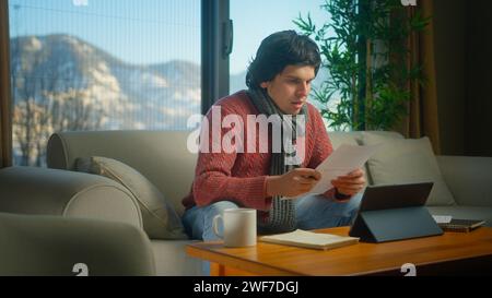 Gli studenti universitari indossano la sciarpa sedersi sul divano per fare ricerche, fare i compiti con il tablet, prendere appunti. Uomo che studia tranquillamente a casa Foto Stock