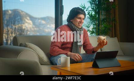 Un uomo sorridente che indossa una sciarpa seduto sul divano mette un paio di monete nel salvadanaio di casa Foto Stock