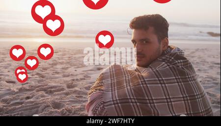 Icone a forma di cuore multiple che fluttuano contro il ritratto di un uomo caucasico con una coperta sorridente sulla spiaggia Foto Stock