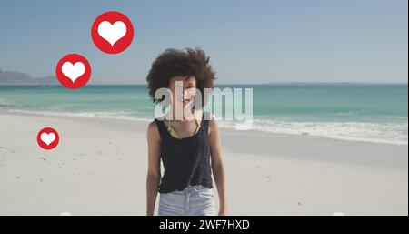 Icone a forma di cuore multiple che fluttuano contro il ritratto di una donna afroamericana sorridente sulla spiaggia Foto Stock