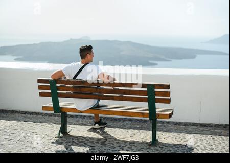 Vista posteriore di un giovane seduto sulla panchina che guarda la costa di Santorini Foto Stock