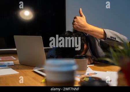 Un uomo d'affari stanco che lavora seduto a dormire ha sdraiato la testa sulla scrivania dell'ufficio con il pc portatile e mostra il gesto del pollice in alto. Foto Stock