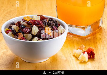 Primo piano di vari tipi di frutta tropicale essiccata in una ciotola di ceramica bianca e succo d'arancia in vetro trasparente su tavola di legno. Foto Stock