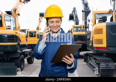 Ingegnere donna in un casco con un tablet digitale in piedi accanto agli escavatori da costruzione. Foto Stock