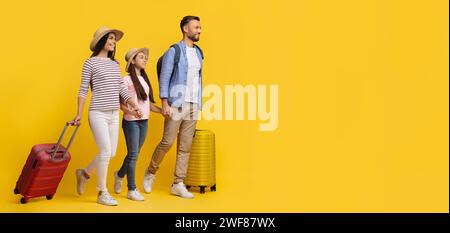 Felice e allegra famiglia caucasica di tre persone che camminano con valigie su sfondo giallo Foto Stock