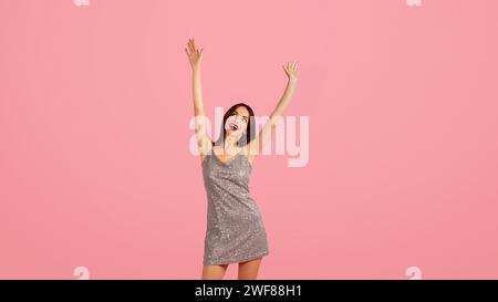 Giovane donna esuberante con un abito in argento scintillante solleva con gioia le mani in alto Foto Stock