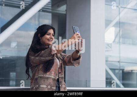 Donna positiva con braccialetto e abiti casual sorridente e con lo smartphone per autoritrarsi contro la costruzione di vetri nelle giornate di sole Foto Stock