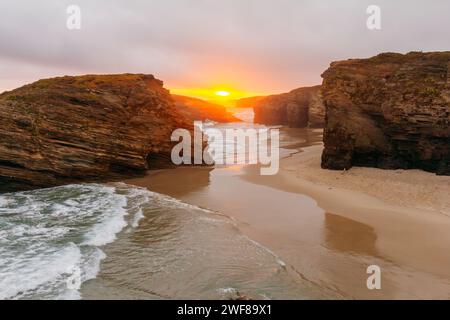 Una tranquilla vista del tramonto su Playa de las Catedrales con onde che si gettano sulla spiaggia di sabbia fiancheggiata da maestose formazioni rocciose. Foto Stock