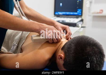 Un operatore sanitario esegue una scansione ecografica sulla schiena del paziente con una sonda ecografica e un gel, con un monitor sullo sfondo Foto Stock