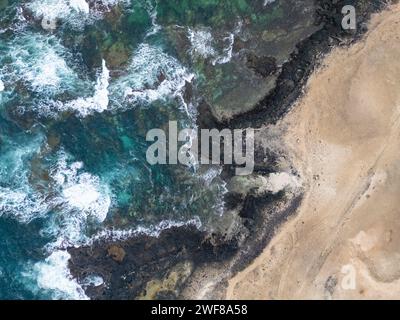 Vista aerea delle onde spumeggianti che si infrangono sulle coste rocciose e sulla spiaggia sabbiosa sulla costa nord di Fuerteventura, vicino a Faro del Toston Foto Stock