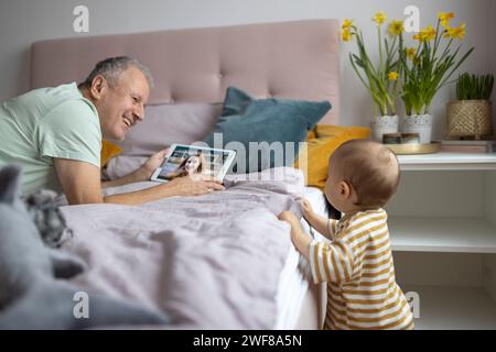Un uomo anziano che parla con una donna su un tablet mentre un bambino interagisce con lui in un ambiente accogliente della camera da letto Foto Stock