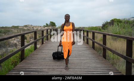 Tutta la donna nera positiva con camice arancione che cammina sul passerella mentre trascina i bagagli e guarda la macchina fotografica contro le montagne e. Foto Stock