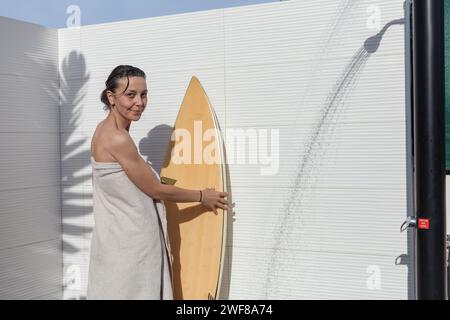 Donna sorridente in un asciugamano che guarda la macchina fotografica accanto a una tavola da surf dopo una doccia Foto Stock