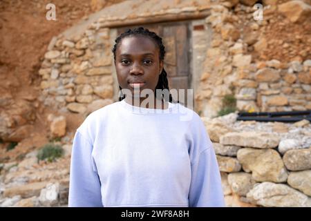 Giovane donna afroamericana sicura di sé che guarda la macchina fotografica davanti a un muro rustico in pietra e a una porta in legno Foto Stock