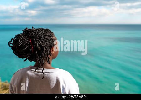 Vista posteriore di una donna afroamericana irriconoscibile e premurosa che guarda tranquillamente lontano dal mare turchese durante il viaggio da solo Foto Stock