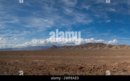 Un'immagine grandangolare che cattura il vasto e arido terreno del deserto di Merzouga in Marocco, con uno sfondo di maestose montagne sotto un cielo blu che spazia Foto Stock