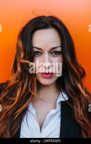 Una giovane donna in posa con camicia bianca e gilet nero regala uno sguardo penetrante con i suoi occhi intriganti, contrastati da un vivace sfondo arancione in Mad Foto Stock