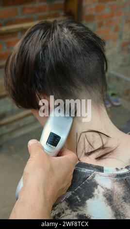 tagliare i capelli di un giovane con lunghi capelli scuri utilizzando un regolacapelli, una vista posteriore, un'immagine verticale, una mano di un uomo tiene un rifinitore con un accessorio e taglia Foto Stock