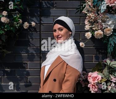 Ritratto di una giovane donna caucasica vestita in un hijab vicino a un muro con fiori. Foto Stock