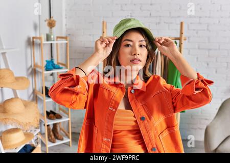 giovane stilista asiatica in abiti arancioni luminosi che indossano un cappello panama e guardano la macchina fotografica in atelier Foto Stock