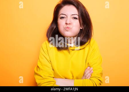 Modello di bellezza sicuro con i capelli brunette che crea un viso baciato sulla fotocamera mentre posa spensierata in studio. Ritratto di una giovane donna adulta con le braccia incrociate che fa un volto goffo. Foto Stock